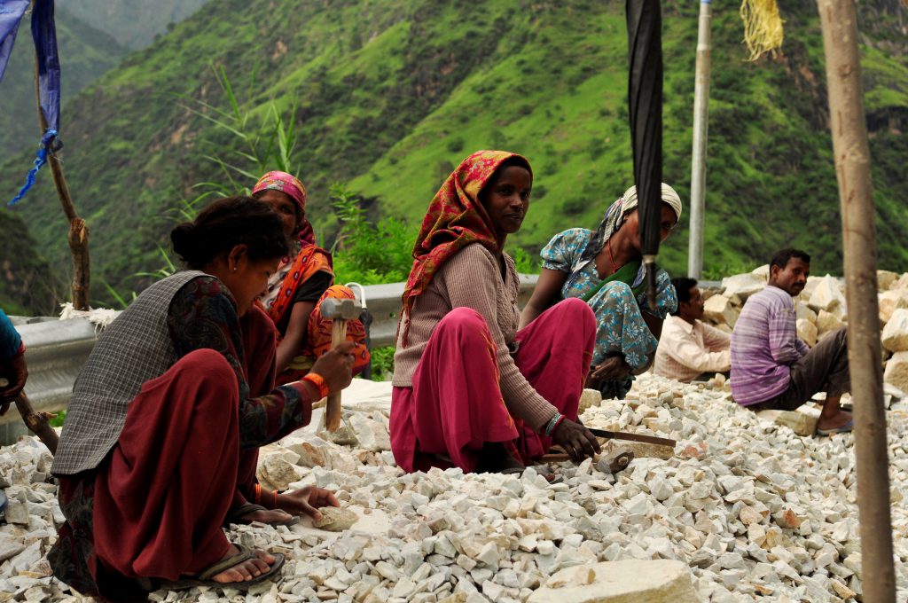 Veibygging i India er håndarbeid. Store steiner knuses med slegge og blir til grus. En spade betjenes av to indere. En liten samling mennesker har tydeligvis ansvar for hver sin lille strekning. 